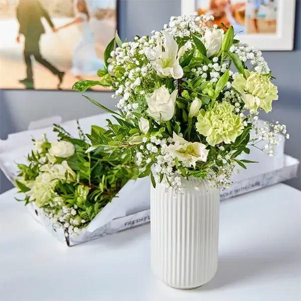A bouquet of fresh flowers in a white ribbed vase, featuring yellow roses, white roses, and clusters of small white daisies with greenery. Behind the vase, an open cardboard box contains additional flowers and foliage, all set against a pink background.